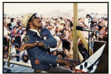 Professor Longhair (1918-1980) performing on an outdoor stage at the 1977 New Orleans Jazz and Heritage Festival. He is seen with his arm raised in the air and a microphone in front of him. A crowd is seen behind him. Professor Longhair performed on Sunday, April 17 at 5:30 p.m. on Stage 1.