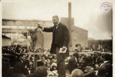 View of an African-American man standing on a platform addressing a crowd of people who surround the platform. He holds a piece of paper in his left hand and points with the index finger of his right hand, his arm uplifted. He wears a long dark coat and trousers that are of a lighter shade. He wears a white shirt, dark tie and weskit. Shows partial view of a watch chain visible through his open coat. Shows buildings in background, papers at his feet.